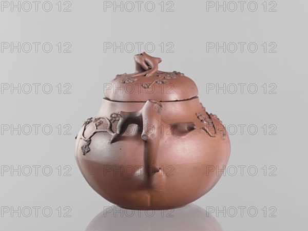 Teapot with a peach-shaped lid, peaches iconography, Chinese ceramics, flowers, trees, twigs, Yixing style. Handle and spout shaped like tree branches with flowering branches growing around the vessel. Red stoneware, 18th century, China. Vessel; height 11.8 cm, width 17.5 cm., A round, reddish-brown ceramic pot with a lid sits against a pale gray background, showing a smooth, slightly glossy surface and a few darker patina spots; the lid has a sculpted handle resembling a twisted organic form and the body features raised vine-like decorative elements and small flower-like motifs in slightly darker brown, with a thick, branch-like handle extending from the top toward the midsection and an uneven, hand-formed rim; the pot rests on a reflective surface that casts a soft mirrored hint of its rounded base.