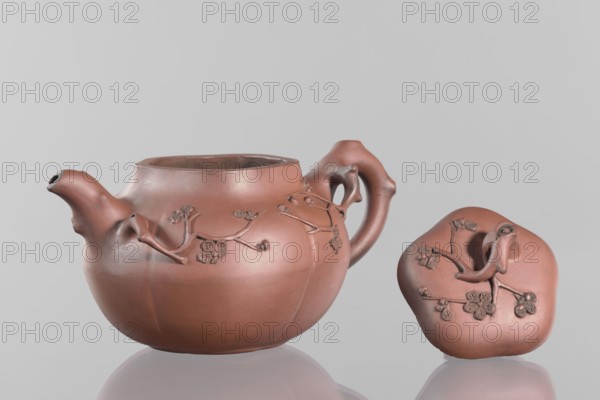 Teapot with a peach-shaped lid;, Red stoneware teapot, Yixing style; handle and spout shaped like tree branches with flowering branches growing around the vessel. Decoration includes peaches, flowers, trees, and twigs. 18th century China. Vessel height 11.8 cm, width 17.5 cm., A reddish-brown ceramic teapot and its matching lid sit on a reflective surface against a light gray background; the teapot has a rounded body with a short curved spout and a sculpted handle, both showing organic, branch-like forms, and both pieces feature raised floral branch decorations in the same reddish-brown tone with slightly darker accents.