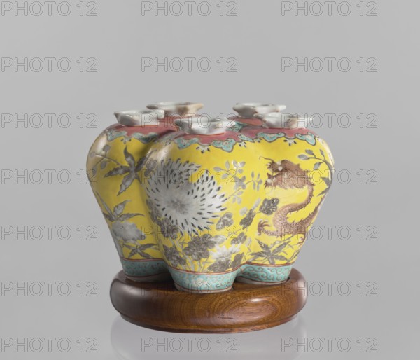 Vase in the shape of five connected vases on a base, Chinese, colored porcelain with dragons and floral decorations, ceramics, 18th/19th century China, vessel, height 15.8 cm, The image shows a ceramic vase with a bulbous, multi-lobed shape resting on a round wooden stand; the vase surface is predominantly bright yellow with painted floral and foliage motifs in muted gray and white, and a stylized dragon or serpentine creature in brownish-red visible on one side; the upper rim area has a band of coral pink with turquoise-green scalloped accents, and the feet or base edges of the vase feature a turquoise-blue patterned border; the wooden stand is warm brown with visible grain, and the background is a plain light gray.