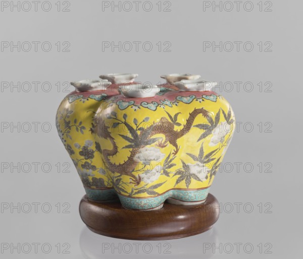 Vase in the shape of five connected vases on a base, Chinese, colored porcelain with dragon and floral decoration, ceramics, 18th–19th century, China, vessel, height 15.8 cm, A ceramic vessel with a bulbous, lobed shape resting on a round wooden stand; the vessel has a bright yellow background with painted floral motifs and a sinuous dragon-like creature in brown, surrounded by leaves and white flowers, a turquoise band with decorative pattern near the base, and a reddish-pink rim area topped by a group of white flared openings; visible colors include yellow, brown, white, turquoise, reddish-pink, gray, and natural wood tones.