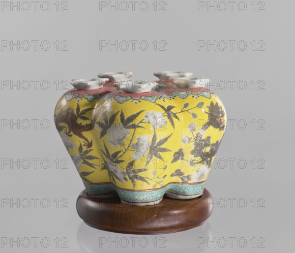 Vase in the shape of five connected vases on a base, Chinese, colored porcelain, dragons and floral decorations, ceramics, 18th/19th century China, vessel, height 15.8 cm, A ceramic vessel with a rounded, lobed shape sits on a wooden stand; the vessel has a bright yellow body decorated with floral and leafy motifs in gray and white, and features ornamental borders in turquoise and pink near the top and bottom. The top edge of the vessel shows a scalloped white rim with several flared openings, and the surface includes painted elements in brown and darker gray that resemble animal or bird forms. The wooden stand is brown with a smooth, polished finish, and the background is a neutral light gray.