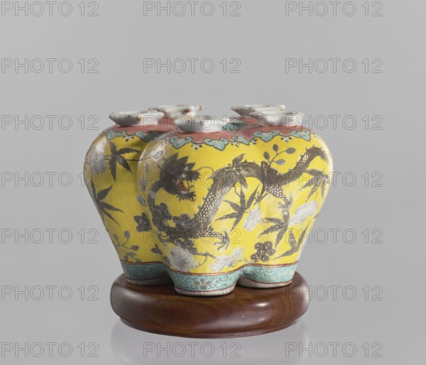 Vase in the shape of five connected vases on a base; maker, Chinese; colored porcelain with dragons and floral decorations; ceramics; ; 18th–19th century, China; vessel; height 15.8 cm, A yellow ceramic object with a glossy finish sits on a round wooden base; the ceramic piece has a bulbous, multi-lobed shape with a scalloped top and several small white rimmed openings, and features painted floral and dragon motifs in dark gray and black with accents of white and muted green; a decorative band in turquoise with a cloud-like pattern runs along the lower edge, and a pinkish border with teal details encircles the top; the wooden base is brown and polished, and the background is a smooth light gray.