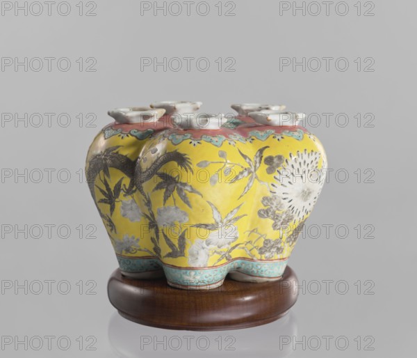 Vase in the shape of five connected vases on a base, Chinese, colored porcelain, dragons, floral decorations, ceramics, 18th/19th century, China, vessel, height 15.8 cm, An ornate ceramic vase with a bulbous, lobed body sits on a wooden stand; the vase has a bright yellow background with decorative floral and foliage motifs painted in shades of gray and white, and a large circular floral burst rendered in white and gray on one side. The top rim area is banded with a pinkish color accented by turquoise-blue scalloped patterns, and the base edge features a turquoise-blue band with a repeating decorative motif. The vase has a group of small, flared white openings at the top. The wooden stand is a rich brown with a smooth finish. Visible colors include yellow, gray, white, pink, turquoise-blue, brown, and a hint of black in the decorative detailing.