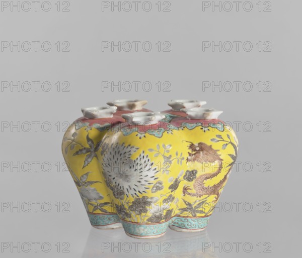 Vase in the shape of five connected vases on a base, Chinese, colored porcelain with dragon and floral decorations, 18th/19th century China, vessel, height 15.8 cm., A ceramic object with a glossy finish sits against a plain light gray background, featuring a bright yellow body decorated with floral and dragon-like motifs in muted grays, browns, and rust tones, and accents of turquoise and pale blue along the lower rim. The top edge is surrounded by a pinkish band with scalloped turquoise detailing, and several small white cup-shaped openings with smooth rims are arranged across the top. The surface shows painted leaves and a large radiating flower in pale gray and white, and a sinuous dragon-like figure in brownish-red on one side. The piece casts a soft reflection on the surface beneath it.