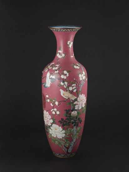 Vase, cloisonné enamel, late 19th century Meiji Period, Japan. Long, narrow neck flaring at the spout; height 123 cm, diameter 38 cm, weight 13.0 kg. Decoration: doves among blooming peonies and cherry trees on a pink background. Subjects: cherries, peonies, doves., A tall, slender vase with a flared rim sits against a dark background, the vase surface showing a dominant deep pink color with decorative floral branches and blossoms in white and pale pink, accompanied by green leaves and darker green foliage. The design includes a couple of birds rendered in soft beige and pale blue tones perched among the branches, and the rim and base feature narrow bands with blue and gold accents. The overall palette includes deep pink, white, pale pink, green, dark green, beige, pale blue, blue, gold, and brown.