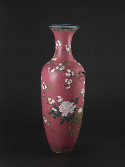 Vase, cloisonné enamel, late 19th century Meiji period, Japan. Long narrow neck flaring at the spout; height 123 cm, diameter 38 cm, weight 13.0 kg. Decoration: doves among blooming peonies and cherry trees on a pink background; motifs include cherries and peonies., A tall, narrow vase with a curved neck and flared rim sits against a dark background; the vase has a deep pink surface decorated with a pattern of white blossoms on thin brown branches and green leaves, with a cluster of larger pale pink flowers and darker green foliage near the middle, a band of blue and gold ornamentation around the rim and base, and a small area of surface damage showing a rough, darker break.