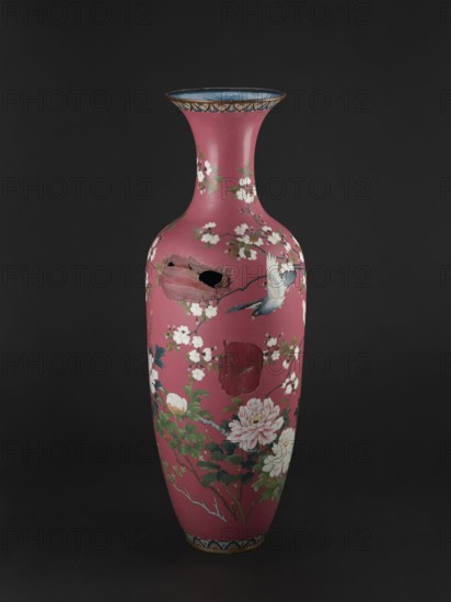 Vase, Japanese, Meiji period late 19th century. Cloisonné enamel vessel with long narrow neck flaring at the spout. Decoration: doves among blooming peonies and cherry trees on a pink background. Height 123 cm; diameter 38 cm; weight 13.0 kg., A tall, narrow vase with a flared rim sits against a dark background; the vase is predominantly deep pink with painted floral and bird motifs in white, green, blue, and touches of yellow and brown, and it shows visible damage with a jagged hole and an area of surface loss exposing darker interior material.