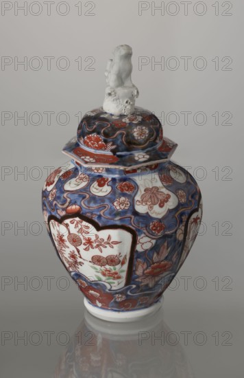 Vase with cover, Imari porcelain, 18th century Edo period, Japan. Decoration: flowers, tree, bamboos, peonies, karashishi mythical lion-dog. Vessel, height 25.5 cm, diameter 16 cm., A lidded porcelain jar with a glossy finish sits against a pale grey background; the jar is predominantly painted in shades of deep blue and red with white areas, and a small white sculpted figure tops the lid. The curved body and flared neck of the jar are covered in swirling blue patterns and red floral motifs, with several white cartouche-shaped panels that contain more red floral decorations and touches of green. The lid echoes the blue and red design with cloud-like shapes, and the white sculpted finial has an uneven, slightly rough texture. The base of the jar is white and reflects faintly on the glossy surface beneath it.