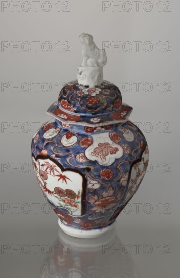 Vase with cover, Japanese, Edo period 18th century, imari porcelain decorated with flowers, tree, bamboos, peonies, and karashishi; vessel, height 25.5 cm, diameter 16 cm., A decorated ceramic jar with a lid topped by a small sculpted figure sits against a neutral gray background. The jar is predominantly blue with areas of white and red-orange, and it shows floral and cloud-like motifs in a painted pattern. Several white panels framed in dark outlines contain additional floral scenes with red-orange and green accents. The lid echoes the blue and red-orange floral design and has a white sculpted figure on top. The jar rests on a white foot and casts a faint reflection on the glossy surface beneath it.