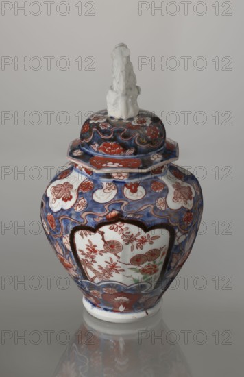Vase with cover, Japanese, Edo period 18th century, Imari porcelain decorated with flowers, bamboo, peonies and karashishi motifs; vessel, height 25.5 cm, diameter 16 cm., A ceramic lidded vase with a sculpted white finial sits against a neutral gray background, the body and lid decorated in a mix of deep blue, red, white, and touches of green and brown forming floral and curvilinear patterns, with scalloped white panels on the body containing red floral motifs, a hexagonal rim where the lid meets the vase, and a glossy surface that reflects onto the smooth surface beneath.