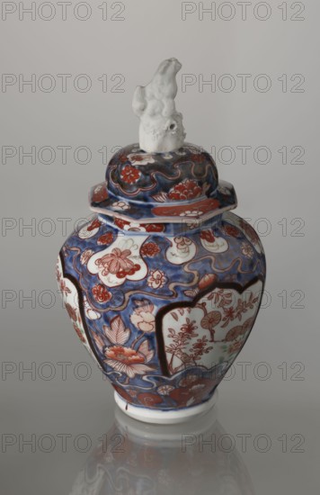 Vase with cover, Japanese, Edo period 18th century; porcelain, Imari style; decorated with flowers, tree, bamboos, peonies, and karashishi mythical lion-dog; vessel; height 25.5 cm, diameter 16 cm., A ceramic lidded jar with a rounded body and a flared neck, topped by a sculpted finial in an off-white color, sits against a neutral gray background and reflects faintly on the surface below; the jar is decorated with flowing cloudlike patterns and floral motifs in deep blue and rich red tones, with areas of pale white where floral scenes are framed by dark outlines, and accents of muted green appearing subtly among the painted blossoms.