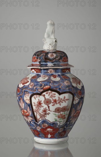 Vase with cover, Japanese ceramics imari, 18th century Edo period, Japan. Portrays flowers, tree, bamboos, peonies, and karashishi; vessel measures height 25.5 cm, diameter 16 cm., A lidded porcelain vase with a sculpted figure on its lid is shown against a neutral gray background; the vase displays a dominant palette of blue, red, white, and touches of green, with ornate floral and cloudlike patterns covering its curved body and lid; the lid has a white sculpted figure with a textured base, and the vase body features white cartouche-shaped panels framed in dark outlines containing red floral motifs, surrounded by flowing blue backgrounds with red and white blossoms and leaf designs, and a white foot and rim.