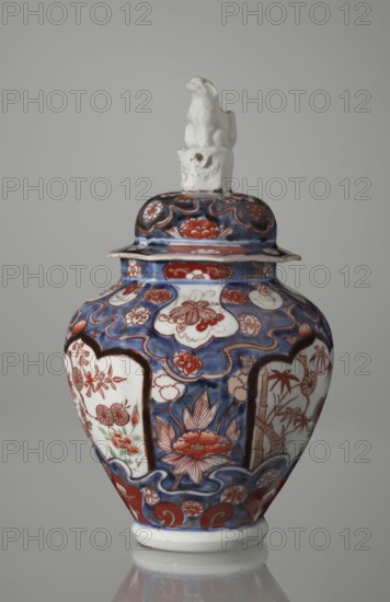 Vase with cover, Japanese, Edo period 18th century, imari ceramics decorated with flowers, tree, bamboos, peonies, and karashishi; vessel, height 25.5 cm, diameter 16 cm., A porcelain lidded jar with a glossy surface sits against a plain gray background; the jar has a rounded body tapering to a narrower base and a lid topped by a small sculpted animal figure in white; the main decorative palette includes deep blue, red, white, and touches of green, with intricate floral and cloud-like motifs painted across the body and lid; panelled areas on the jar display stylized flowers and foliage outlined in red against white, while surrounding sections are filled with swirling blue backgrounds and red floral accents; the rim and foot are edged in white, and the overall finish is smooth and reflective.