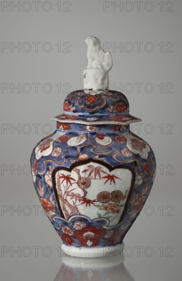 Vase with cover, Japanese, Edo period 18th century, imari porcelain vessel decorated with flowers, tree motifs, bamboos, peonies, and karashishi; height 25.5 cm, diameter 16 cm., A lidded porcelain jar with a glossy finish sits against a neutral gray background, predominantly decorated in deep blue and rich red with areas of white and small touches of green and gold. The lid is topped by a white sculpted figure. The body of the jar features ornate floral and wave-like patterns in blue and red, with a white panel on the front containing a botanical scene rendered mainly in red and accented with green and pale gold. The rim and base have banded decorative motifs in the same blue and red palette, and the jar's curved silhouette is reflected faintly on the surface beneath it.