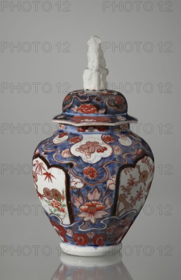 Vase with cover, Japanese, Edo period 1603–1868, 18th century; porcelain imari vessel decorated with flowers, tree, bamboos, peonies, and karashishi; height 25.5 cm, diameter 16 cm., A ceramic lidded vase with a rounded body sits against a plain light gray background; the lid is topped by a small sculpted figure in white, and the vase surface features ornate painted decoration in deep blue, red, white, and touches of green and brown, with floral motifs, cloud-like cartouches, and flowing scroll patterns covering the body and lid.