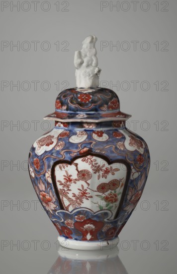 Vase with cover, Japanese, Edo period 18th century, imari ceramics decorated with flowers, tree, bamboos, peonies and karashishi lion-dog iconography; vessel, height 25.5 cm, diameter 16 cm., A lidded porcelain jar with a sculpted finial sits against a plain gray background; the finial is white and depicts a small figure with a textured surface. The jar body is bulbous with a flared rim on the lid and is decorated with an overall palette of blue, red, white, and touches of green; the dominant background decorations are blue with red floral motifs and white cartouche-shaped panels that contain red floral designs and small green accents. The surface shows swirling blue patterns, red blossoms, and stylized leaves, with a band of red and blue ornamental motifs near the base and lid, and the jar rests on a white foot that reflects faintly on the surface below.