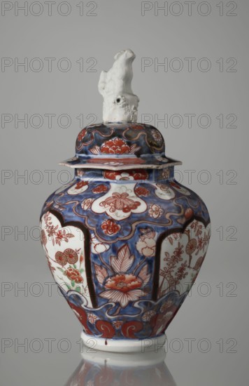 Vase with cover, Japanese, Edo period 18th century, Imari porcelain vessel decorated with flowers, tree, bamboos, peonies, and karashishi; height 25.5 cm, diameter 16 cm., An ornate ceramic lidded jar with a glossy finish sits against a plain gray background, featuring a white sculpted finial on the lid shaped like an abstract animal form; the body of the jar displays a rich palette of deep blue, red, white, and touches of green and brown in a repeating floral and cloud-like pattern, with stylized blossoms, leaves, and curved decorative bands wrapping around its rounded silhouette, and a white base that reflects subtly on the surface below.