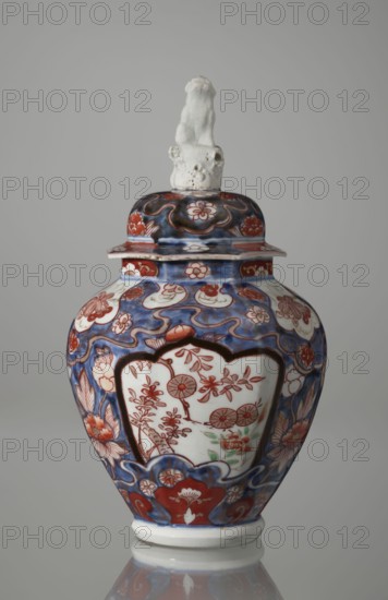 Vase with cover, Japanese, Edo period 18th century, imari ceramics decorated with flowers, tree, bamboos, peonies, and karashishi; vessel, height 25.5 cm, diameter 16 cm., A lidded ceramic vase with a glossy finish sits against a neutral gray background, featuring a predominantly blue and red painted surface with white areas left unpainted; the body is bulbous with a narrow base and a flared rim supporting a domed lid topped by a small white sculpted figure. The painted decoration includes flowing blue waves and cloudlike shapes, red floral motifs and stylized petals, and white panels on the body containing red botanical designs with touches of green. The lid echoes the blue and red floral patterning, and the small sculpted finial is textured and off-white. The base and rim show unpainted white ceramic, and the overall color palette visible is blue, red, white, green, and off-white.