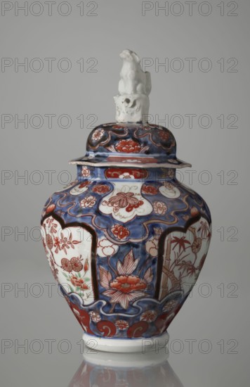Vase with cover, Japanese, Edo period 18th century, Imari porcelain vessel decorated with flowers, tree, bamboos, peonies, and karashishi; height 25.5 cm, diameter 16 cm., A ceramic lidded jar with a glossy finish sits against a plain gray background, topped by a white sculpted finial shaped like a seated animal. The body and lid of the jar display an ornate painted pattern featuring a dominant deep blue background with areas of white that serve as panels for floral and botanical motifs in shades of red and orange, with small touches of green. The decorative design includes stylized flowers and leaves outlined in darker tones, curved wave-like blue borders, and rounded medallion shapes. The jar has a flared rim beneath the lid and a rounded shoulder tapering to a slightly raised white base, and the surface shows subtle reflections from the lighting.
