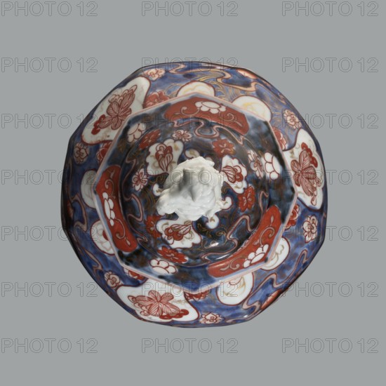 Vase with cover, Japanese, Edo period 18th century, ceramics Imari, decorated with flowers, tree, bamboos, peonies, and karashishi; vessel; height 25.5 cm, diameter 16 cm., A decorative lidded bowl shown from above with a central white sculpted finial; the bowl has a scalloped rim and an inner raised section, both covered in ornate painted patterns featuring floral and cloud-like motifs. The visible colors include blue, red, white, and touches of gold, with darker navy and lighter blue areas blending together; the red appears in panel-like shapes and floral outlines, and white is used for background shapes and the central finial. The overall surface shows glossy ceramic glazing with intricate repetitive designs.