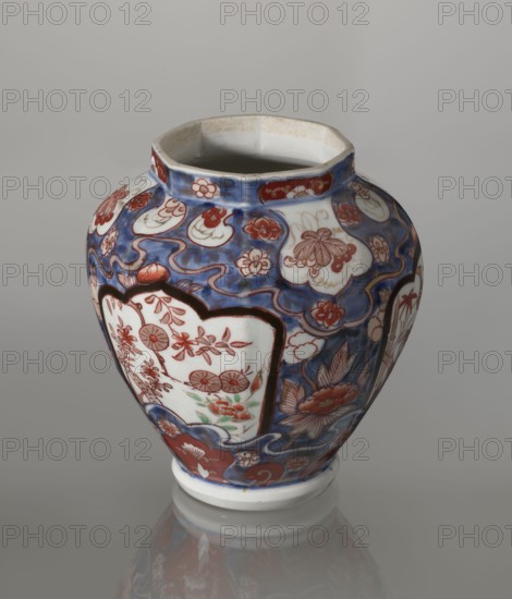 Vase with cover, Japanese, Edo period 18th century, imari porcelain vessel decorated with flowers, tree, bamboos, peonies and karashishi; height 25.5 cm, diameter 16 cm., A rounded ceramic vase with an open top sits on a reflective surface, decorated with flowing blue background patterns and panels outlined in dark tones. The vase shows floral and leafy motifs painted in red and white within scalloped and cloud-like shapes, and there are accents of green among some floral elements. The rim and base are primarily white with slight discoloration at the inner rim, and the overall visible colors include blue, red, white, green, and dark brown.