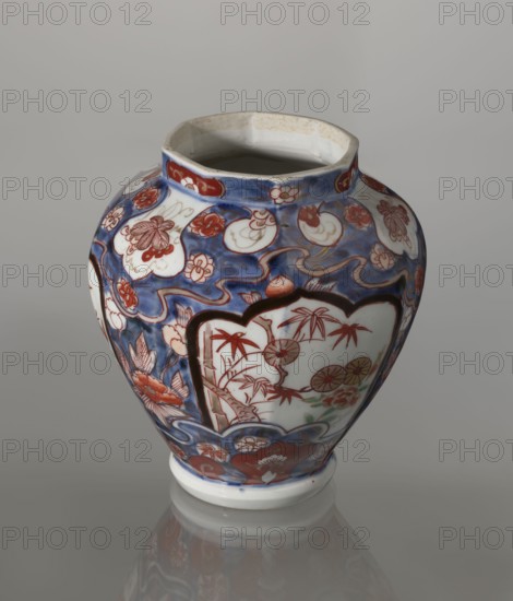 Vase with cover, Japanese, Edo period 1603–1868, 18th century; porcelain imari, decorated with flowers, tree, bamboos, peonies, and karashishi; vessel; height 25.5 cm, diameter 16 cm., A rounded ceramic vase with a wide mouth and a short foot, decorated with a complex painted pattern in blue, red, white, black, green, and brown. The background features deep blue areas with flowing wave-like shapes and scattered white cartouche shapes filled with red floral motifs. A prominent white panel outlined in black on the front shows red-painted bamboo and floral elements with small touches of green and yellow. Around the base and shoulder are red flower designs and stylized decorative borders in blue and red. The mouth interior appears off-white with some brown speckling, and the vase sits on a reflective grey surface against a neutral grey background.