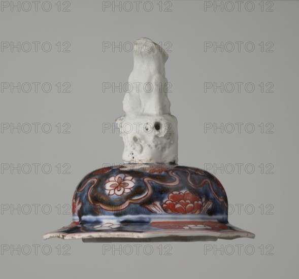 Vase with cover, Japanese, Edo period 18th century, imari ceramics depicting flowers, tree, bamboos, peonies, and karashishi; vessel; height 25.5 cm, diameter 16 cm., A ceramic object with a glossy white sculpted top resembling an irregular organic form sits above a rounded decorative section; the upper portion is matte white with some small pits and rough texture, while the lower rounded area displays a glossy patterned surface in deep blue and red with floral motifs and curving lines, and a flared rim showing white and red elements and some worn edges.