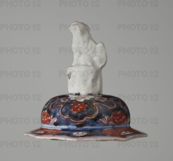 Vase with cover, Japanese ceramics, imari decoration of flowers, tree, bamboos, peonies, and karashishi; 18th century Edo period, Japan; vessel, height 25.5 cm, diameter 16 cm., A ceramic object with a glossy white sculpted figure perched on top of a rounded lid, the figure showing textured fur and a curled tail; the lid below is decorated with floral and curving motifs in deep blue and red with areas of white and small touches of gold, and the rim flares outward into a scalloped edge showing red and white painted panels against a subtle blue background.