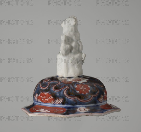 Vase with cover, Japanese, Edo period 18th century, Imari porcelain vessel with decoration of flowers, bamboo, peonies, and karashishi; height 25.5 cm, diameter 16 cm., A ceramic object with a glossy patterned base and a sculpted white form rising from its center; the base shows a deep blue background with flowing wave-like decorative lines and floral shapes in red and white, and areas of dark brown or black accents, while the rim has a band of red with white motifs and a pale underside; the sculpted top is white with a matte glaze, featuring organic, rounded contours and textured details that suggest layered forms.
