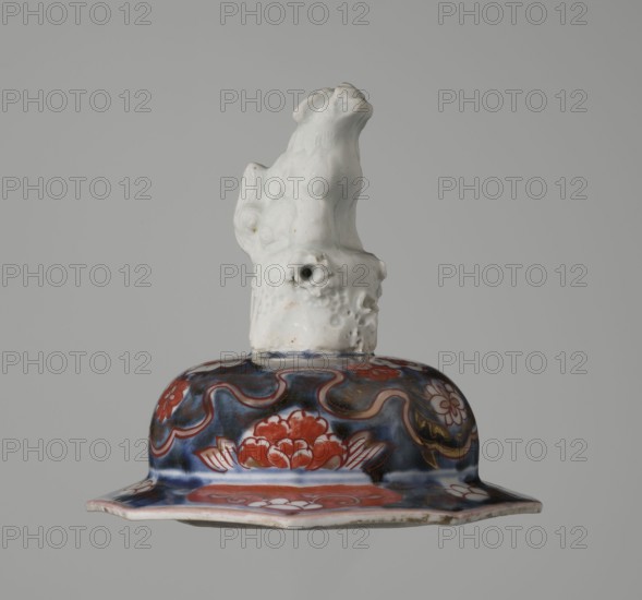 Vase with cover, Japanese, Edo period 18th century, Imari porcelain vessel decorated with flowers, trees, bamboos, peonies, and karashishi; height 25.5 cm, diameter 16 cm., An object resembling a decorative lid with a sculpted animal figure on top, the upper figure is glossy white with textured surface details, and the lower flared section is painted with a dark blue background and red floral motifs accented with gold and white, the rim showing a pale off-white edge and some subtle discoloration.