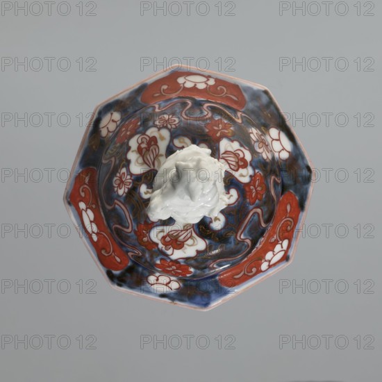 Vase with cover, Japanese, imari porcelain decorated with flowers, tree, bamboos, peonies, and karashishi; Edo period, 18th century; vessel; height 25.5 cm, diameter 16 cm., A top-down view of an octagonal ceramic bowl with a glossy surface showing a central white sculpted element surrounded by decorative floral and cloud-like motifs in red, white, and blue, with areas of dark blue and hints of browning on the glaze.
