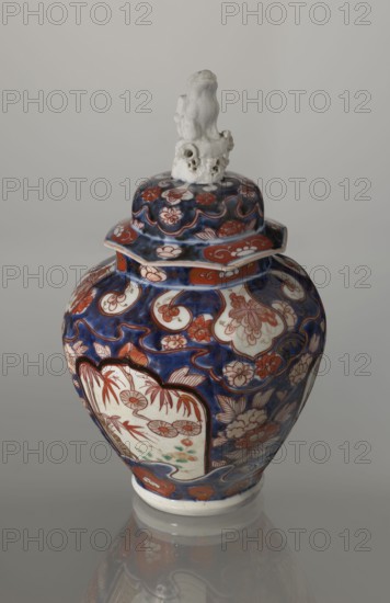 Vase with cover, Japanese, Edo period 18th century, porcelain Imari, decorated with flowers, tree, bamboos, peonies, and karashishi; vessel, height 25.5 cm, diameter 16 cm., A ceramic jar with a lid sits against a light gray background, the jar showing a glossy surface with deep blue, red, white, and touches of green colors; the body is decorated with floral and cloud-like motifs in red and white set against a blue ground, and there are panel-like white areas containing red floral sketches and small green accents; the lid echoes the same blue and red floral patterns and is topped with a sculpted white finial shaped like an abstract organic form with irregular openings and textured surfaces; the jar rests on a white foot and its lower portion and reflection are visible on the smooth surface beneath.