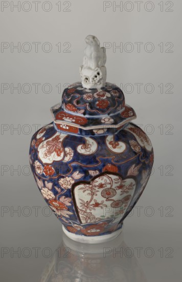 Vase with cover, Japanese, Edo period 18th century, imari porcelain vessel decorated with flowers, tree, bamboos, peonies and karashishi motifs; height 25.5 cm, diameter 16 cm., An ornate lidded ceramic jar with a glossy finish sits against a neutral gray background, featuring a dominant deep blue base color with prominent red and white floral and scroll motifs across its rounded body and lid, a few areas of pale green visible within decorative panels, and a white sculpted figure mounted on the top of the lid; the jar shows intricate painted flowers and curved shapes in red and white set against the blue, with the white base rim and a soft reflection visible on the surface beneath.