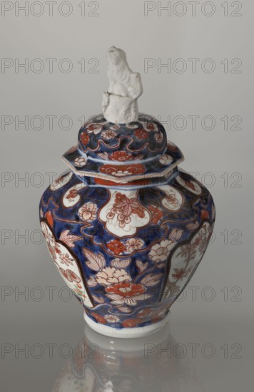 Vase with cover, Japanese, Edo period 18th century, imari ceramics decorated with flowers, tree, bamboos, peonies and karashishi; vessel, height 25.5 cm, diameter 16 cm., A ceramic lidded vase with a glossy surface sits against a plain light gray background, the body and lid covered in an intricate floral pattern in deep blue and reddish-orange with areas of white showing through; the lid is topped by a small white sculpted figure. The vase has a rounded shoulder that tapers toward the base, and the decoration includes stylized flowers and leaves, curving outlines, and panel-like white medallions framed by the blue and reddish-orange motifs. The overall color palette visible is deep blue, reddish-orange, white, and small touches of pale green, with subtle reflections of the vase visible on the glossy surface beneath it.