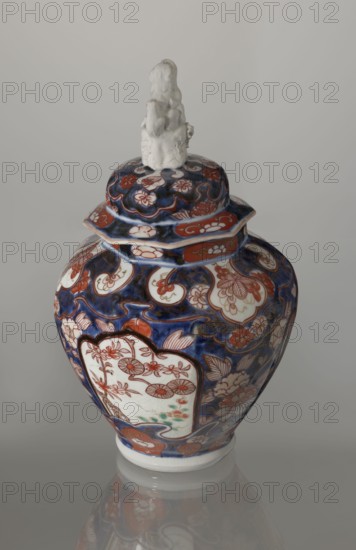 Vase with cover, Japanese Imari ceramics, 18th century Edo period, Japan. Decoration: flowers, tree, bamboos, peonies, karashishi. Vessel, height 25.5 cm, diameter 16 cm., A ceramic covered jar with a glossy finish sits against a neutral pale gray background, featuring a domed lid topped by a small white sculpted figure. The jar displays a rich palette of deep blue, red, white, and touches of green, with intricate floral and cloud-like painted patterns wrapping its curved body and lid. The main body has panels of white framed by red outlines containing stylized flower motifs, set against a dominant cobalt blue field filled with swirling red and white floral designs and goldish accents. The lid echoes the same colors and motifs, and the jar rests on a short white foot that casts a soft reflection on the reflective surface beneath.