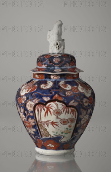 Vase with cover, Japanese, Edo period 18th century, imari ceramics, decorated with flowers, tree, bamboos, peonies and karashishi motifs; vessel; height 25.5 cm, diameter 16 cm., A ceramic jar with a lid topped by a sculpted white finial sits against a plain gray background; the jar's surface shows a deep blue base color with red and white decorative motifs, including floral shapes and panels with red-painted bamboo and circular flower-like designs, along with green accents near the base of the painted panels, and the lid and body feature repeating red and white floral patterns on the blue field.