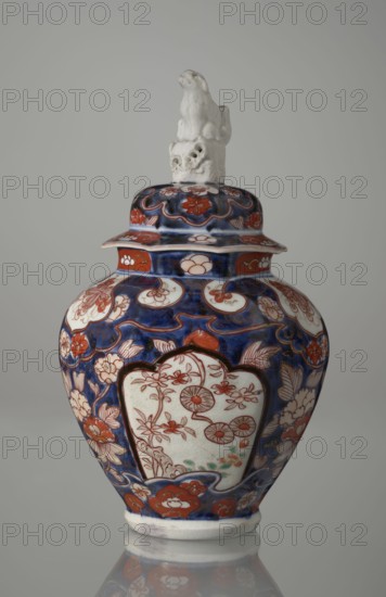 Vase with cover, Japanese, Edo period 18th century, imari ceramics depicting flowers, tree, bamboos, peonies and karashishi; vessel, height 25.5 cm, diameter 16 cm., A lidded ceramic vase with a glossy finish sits against a plain neutral background; the vase has a rounded body that narrows toward the base and a lid topped with a small sculpted animal figure in an off-white color. The main surface decoration features a deep blue ground with a pattern of floral and foliate motifs in shades of red and off-white, with some small areas of pale green visible in the floral panels; the lid echoes the same blue and red floral design. The body displays several off-white cartouche-shaped panels outlined in dark color containing delicate red floral linework, and the lower rim shows a band of red floral forms on the blue background. The sculpted finial on the lid is textured and off-white, and the vase rests on a short off-white foot that reflects slightly on the glossy surface beneath.
