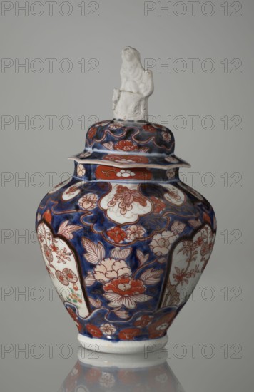 Vase with cover, Japanese, Edo period 18th century, imari porcelain vessel decorated with flowers, tree, bamboos, peonies, and karashishi karajishi. Height 25.5 cm; diameter 16 cm., A decorative lidded vase with a glossy surface sits against a plain neutral background, featuring a deep blue base color with prominent red and white floral and cloud-like patterns across its curved body and lid; the vase shows panels with lighter white areas containing detailed red floral motifs, and areas of darker blue filled with red and white blossoms and leaves; the rounded lid is topped by a small sculpted figure in an off-white color with a slightly textured surface; the overall palette includes blue, red, white, and subtle touches of green, with a reflective sheen visible on the vase and a faint reflection beneath it.