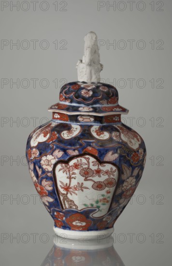 Vase with cover;, Japanese, Edo period 18th century; porcelain, Imari style; decoration: flowers, tree, bamboo, peonies, karashishi; vessel; height 25.5 cm, diameter 16 cm., The image shows a decorative lidded vase with a sculpted finial on top, the finial appearing white and textured. The vase body and lid feature an intricate floral and ornamental pattern in deep blue and reddish-orange with accents of white and small areas of green. The vase has rounded shoulders tapering toward a narrower base, and the patterns include stylized flowers and curved motifs. The background is a plain light gray, and the vase sits on a reflective surface that shows a faint mirror image of the base.