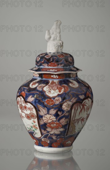 Vase with cover, Japanese, Edo period 18th century, imari porcelain vessel decorated with flowers, tree, bamboos, peonies, and karashishi; height 25.5 cm, diameter 16 cm., A lidded ceramic vase with a glossy finish sits against a plain gray background, the body of the vase showing a deep cobalt blue base color with decorative floral and foliate motifs in reddish-orange and white, along with accents of green and pale brown; the overall shape is bulbous with a flared rim and a short foot that rests on a reflective surface creating a subtle mirror image. The lid repeats the blue and reddish-orange floral patterns and is topped by an off-white sculpted finial with an irregular, organic form and a slightly rough texture. The decoration includes panel-like white cartouches filled with reddish-orange floral designs and surrounding blue areas populated by stylized blossoms and leaves, with thin dark outlines emphasizing shapes and contours.