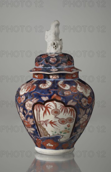 Vase with cover, Japanese, Edo period 18th century, imari ceramics depicting flowers, bamboo, peonies, and karashishi; vessel, height 25.5 cm, diameter 16 cm., A ceramic lidded jar with a glossy surface, featuring a deep blue background with extensive red and white floral and cloud-like motifs, and accents of green and brown in the painted panels; the jar has a bulbous body tapering to a narrow base with a white foot, a flaring rim under the lid, and a lid topped by an irregular white sculpted finial that has a rough, textured appearance; the painted decoration includes framed panels on the body with stylized botanical scenes in red and brown on a white ground, surrounded by red and white flowers and swirling blue patterns, and the overall color palette visible is blue, red, white, green, brown, and a hint of black in the outlining.