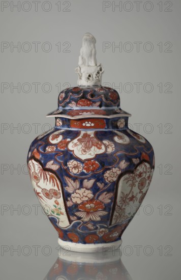 Vase with cover, Japanese, Edo period 18th century, Imari ceramics, decorated with flowers, tree, bamboos, peonies, and karashishi; vessel; height 25.5 cm, diameter 16 cm., A ceramic lidded vase with a glossy finish stands against a neutral gray background, featuring a predominantly deep blue surface decorated with floral motifs in shades of red, white, and pale green, and bordered areas of off-white that contain red floral line drawings; the body has curving panels and scalloped borders in blue and red, while the lid repeats the blue and red floral patterns and is topped by a white sculpted finial with an irregular, slightly textured form.