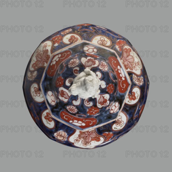 Vase with cover, maker, Japanese Imari porcelain vessel depicting flowers, tree, bamboos, peonies, and karashishi; ceramics, Edo period 18th century, Japan; height 25.5 cm, diameter 16 cm., A round ceramic object with a scalloped rim viewed from above, predominantly showing blue, white, and red colors with gold accents; the surface features a repeating floral and cloud-like pattern in red and white set against deep blue areas, with several white panels containing red floral linework; a central raised white sculpted element sits atop the patterned interior; the background behind the object is a flat gray.