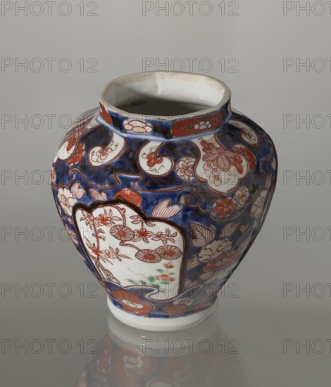 Vase with cover, Japanese, Edo period 18th century, imari porcelain vessel decorated with flowers, tree, bamboos, peonies and karashishi; height 25.5 cm, diameter 16 cm., A ceramic vase with a rounded body and a narrow neck sits on a reflective surface, showing a reflection beneath it; the vase is predominantly deep blue with areas of white and decorated with red-orange floral and cloud-like motifs and goldish accent lines, with a few greenish touches visible within a framed white panel on the body; the neck interior is off-white with some beige discoloration, and the rim is slightly irregular.