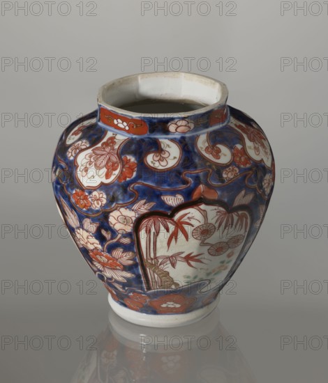 Vase with cover, Japanese, Edo 1603–1868, 18th century, imari ceramics depicting flowers, tree, bamboos, peonies, and karashishi; vessel; height 25.5 cm, diameter 16 cm., A rounded ceramic vase with a wide mouth and a flared rim sits against a neutral gray background, decorated with a deep blue ground and intricate floral and botanical motifs in red, white, and touches of green, with accents of brownish-gold; the design includes stylized flowers, leaves, and framed panels with botanical scenes, and the vase rests on a white foot with a subtle reflected image on the glossy surface below.