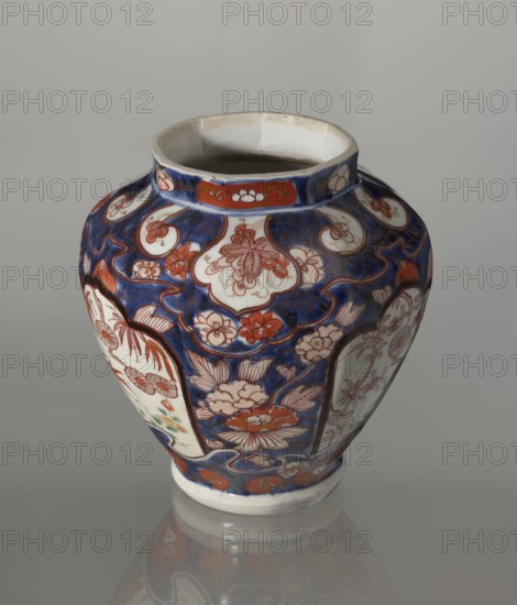 Vase with cover, Japanese, Edo period 18th century, imari porcelain vessel decorated with flowers, tree, bamboos, peonies, and karashishi; height 25.5 cm, diameter 16 cm., A rounded ceramic vase with a wide mouth sits against a neutral gray background, decorated with a deep blue ground and a pattern of floral shapes and curved panels in shades of red, white, and a touch of green. The vase shows several stylized flowers and leaves in red and white set within blue areas, with white panels bordered by red outlines that contain more floral designs. The rim and base are white with some red decorative elements near the top band, and the overall surface has a glossy finish that creates a soft reflection on the surface beneath it.