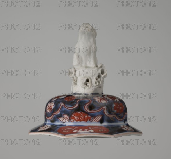 Vase with cover, Japanese, Edo period 18th century, imari ceramics depicting flowers, tree, bamboos, peonies, and karashishi; vessel, height 25.5 cm, diameter 16 cm., A ceramic object with a glossy white sculpted top resembling an abstract organic form sitting on a decorative flared base; the base shows a dark blue background with a few red floral and wave-like motifs and touches of white highlights, and the overall image background is a uniform light gray.