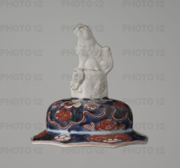 Vase with cover, Japanese, Edo 1603–1868, 18th century, imari ceramics depicting flowers, tree, bamboos, peonies, and karashishi; vessel, height 25.5 cm, diameter 16 cm., The image shows a ceramic object with a sculpted white figure of an animal, possibly a dog or lion, perched on a white textured pedestal atop a decorative base. The base features a rich dark blue background with reddish-orange floral and wave-like patterns and some white accents. The rim of the base is scalloped and shows pale pinkish edges. The overall background of the photograph is a plain light gray.