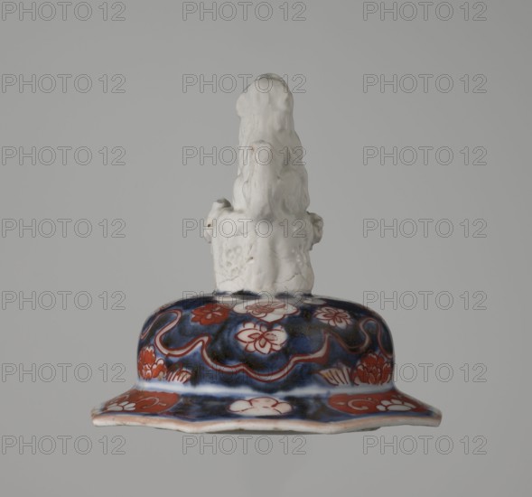 Vase with cover, Japanese ceramics imari, decorated with flowers, tree, bamboos, peonies, and karashishi; 18th century Edo period, Japan; vessel; height 25.5 cm, diameter 16 cm., An object with a glossy white sculpted top resembling a stylized figure or form sits atop a domed base decorated with a dark blue background and reddish-orange floral and wave-like motifs, with areas of white showing through the painted patterns; the base has a flared, slightly undulating rim and the overall color palette includes white, dark blue, reddish-orange, and small touches of pale beige.