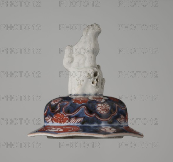 Vase with cover, Japanese, Edo period 18th century, 1603–1868, imari ceramics; decorated with flowers, tree, bamboos, peonies, karashishi; vessel; height 25.5 cm, diameter 16 cm., A ceramic object with a glossy white sculpted top resembling an abstract animal or figure with textured details and small holes sits atop a rounded, flared base. The base is decorated with a pattern of floral and wave-like motifs in deep blue and rich red on a white background, with areas of darker shading and highlights that give a glazed appearance. The overall color palette includes white, deep blue, red, and touches of darker tones against a plain light gray background.